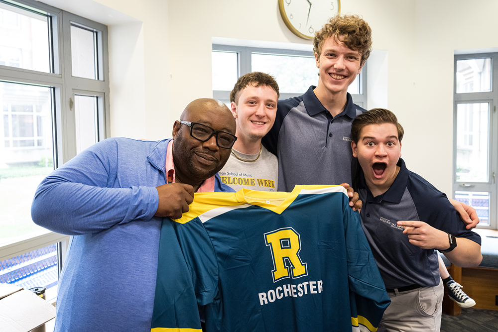 John Blackshear holds a URochester jersey while a group of students cheers behind him..